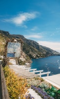 A scenic coastal view featuring a rustic sign for a restaurant with lush greenery and flowers in the foreground. The sign reads 'Ristorante il Capitano'. The landscape displays a beautiful cliffside town with buildings scattered along the cliffs overlooking a tranquil blue sea. Numerous small boats dot the water, and the sky is a clear blue with a few white clouds.