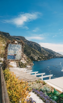 A scenic coastal view featuring a rustic sign for a restaurant with lush greenery and flowers in the foreground. The sign reads 'Ristorante il Capitano'. The landscape displays a beautiful cliffside town with buildings scattered along the cliffs overlooking a tranquil blue sea. Numerous small boats dot the water, and the sky is a clear blue with a few white clouds.