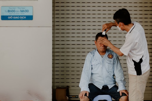 A man sitting on a stool receives a haircut from another man. The barber is wearing a face mask and trims the recipient's hair using clippers while gently holding his head steady. The setting appears to be an informal outdoor or street setup, with a closed metal shutter in the background.