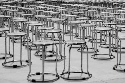 A large number of metal stools are arranged in a repetitive pattern on a smooth, light-colored surface. The rows of stools create a sense of symmetry and order, with each stool composed of a circular seat and simple metal legs. The image appears to be captured in grayscale, emphasizing texture and form.