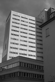 A tall, rectangular office building features a grid of windows, with an adjacent lower structure in the foreground. The overall aesthetic is modernist, characterized by clean lines and a lack of ornamentation. A large advertisement is displayed on one side of the building.