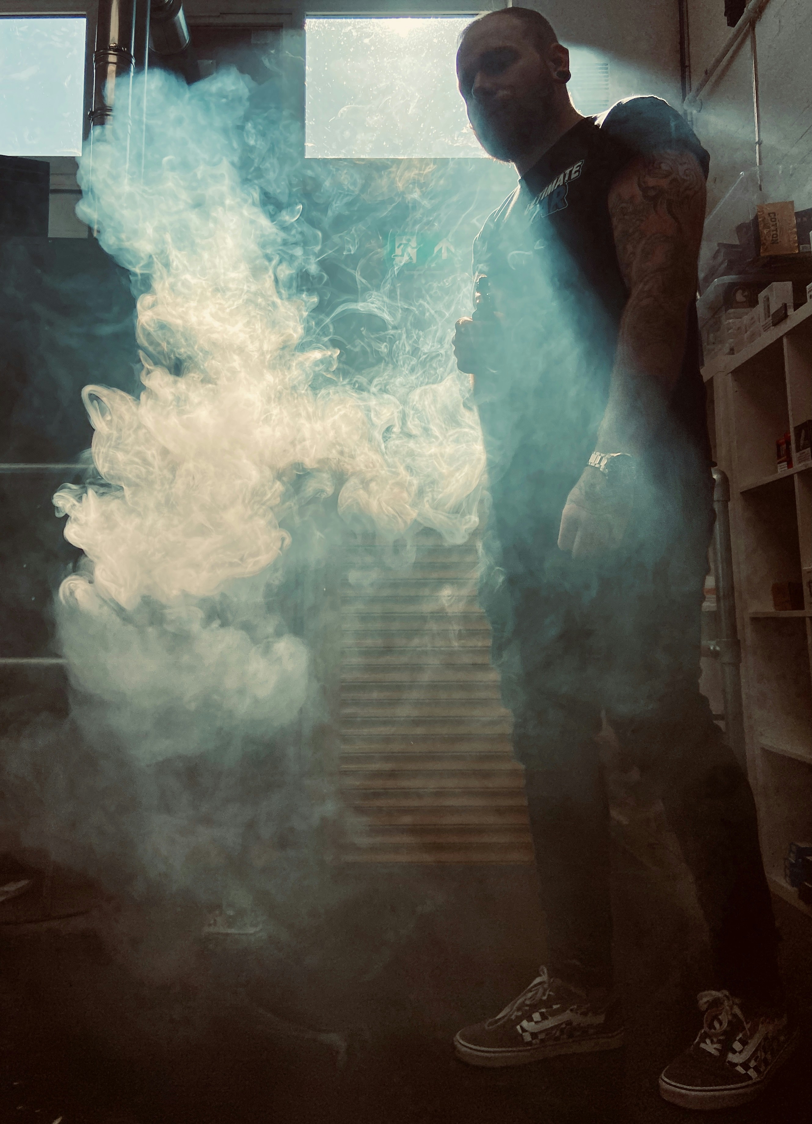 A tall handsome man with a dusty black shirt vaping his ultimate puff eliquid juice in silhouette and moody warehouse