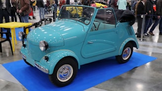 A turquoise, vintage-style microcar is parked on a blue mat in an indoor exhibition space, surrounded by people. The car is compact with a convertible roof, round headlights, and a retro design. Nearby, there are some black and yellow chairs around a small table.