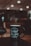 A black coffee mug featuring the motivational phrase 'Always Do What You Love' and the brand name 'wework' in white text. The mug is set on a dark surface with a blurred background that includes warm, ambient lighting from ceiling lights.