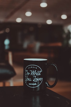 A black coffee mug featuring the motivational phrase 'Always Do What You Love' and the brand name 'wework' in white text. The mug is set on a dark surface with a blurred background that includes warm, ambient lighting from ceiling lights.
