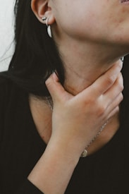 A person with a hand placed on their neck, wearing a black shirt. The person has a silver necklace and a white earring. The skin tone is slightly warm, with natural textures visible.