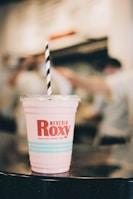 A pink beverage in a plastic cup with a logo and the name 'Neveria Roxy' printed on it, featuring a striped black and white straw. The background is softly blurred, indicating a busy indoor setting.