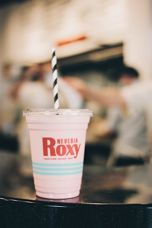 A pink beverage in a plastic cup with a logo and the name 'Neveria Roxy' printed on it, featuring a striped black and white straw. The background is softly blurred, indicating a busy indoor setting.