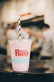 A pink beverage in a plastic cup with a logo and the name 'Neveria Roxy' printed on it, featuring a striped black and white straw. The background is softly blurred, indicating a busy indoor setting.