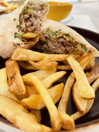A plate of crispy golden-brown fries is served alongside a wrap filled with shredded meat, lettuce, and possibly other vegetables. The meal is arranged on a dark plate with a bowl containing a yellow sauce in the background.