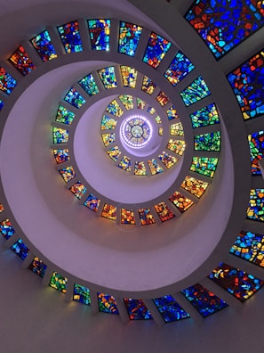 A spiral staircase composed of vibrant stained glass panels in various colors, creating a dynamic and colorful tunnel effect. The light filtering through the glass casts an array of colorful reflections within the spiral structure.