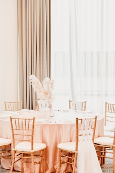 A round table is elegantly set with a pink tablecloth and surrounded by gold chiavari chairs. In the center of the table, there is a vase with dried pampas grass as the centerpiece. The setting is well-lit by natural light coming through large sheer curtains in the background, creating a soft and calming atmosphere.