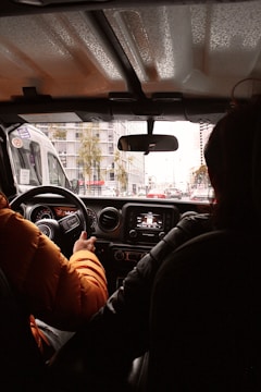 The view from the backseat of a car, showing two people sitting in the front seats. The driver, wearing an orange jacket, is holding the steering wheel. The dashboard and controls are visible. The car is in a city setting with tall buildings and traffic visible through the windshield.