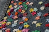 A close-up view of denim fabric adorned with an array of small, colorful embroidered flowers. The flowers vary in shades of red, orange, yellow, green, blue, pink, and purple, creating a vibrant and cheerful pattern across the fabric.