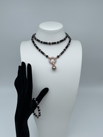 A mannequin bust wearing a black and gold beaded necklace with a large round pendant, paired with a matching beaded bracelet on a black velvet glove.