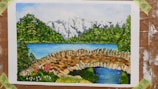 A colorful painting depicts a serene landscape with a stone bridge over a small stream in the foreground. In the middle ground, there's a vibrant blue lake surrounded by lush green trees. Snow-capped mountains are visible in the background under a bright sky.