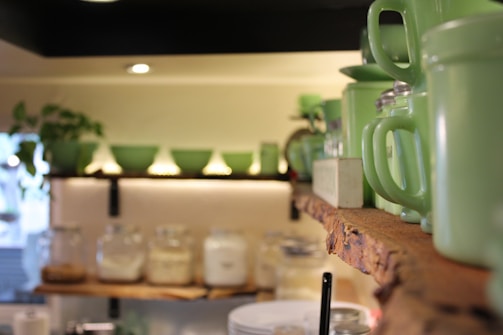 A cozy kitchen or pantry shelf featuring a collection of vintage green ceramic dishes, cups, and jars. The background includes jars filled with various ingredients, a warmly lit space, and a small plant adding a touch of greenery.