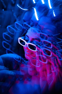 A visually striking image features a woman wearing white-framed sunglasses amidst a background of vibrant neon lights. The effect creates multiple overlapping reflections or projections of her face, contributing to a psychedelic and dynamic composition.