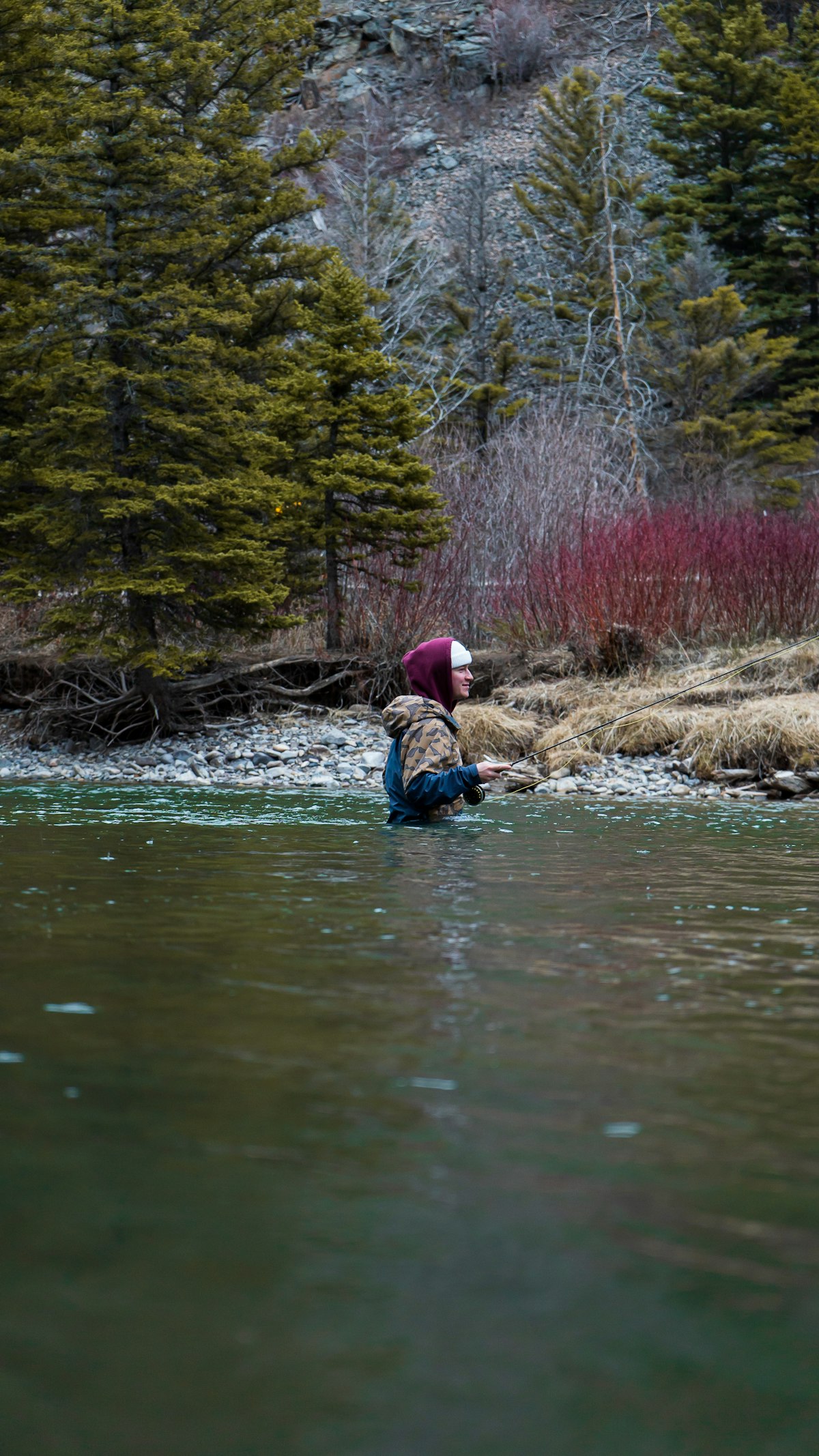 A fly angler waist-deep in cold, opaque green water, rod held out to the side, spruce and red-stemmed willow on the bank behind — the kind of water where you cannot see the bottom and have to read it another way