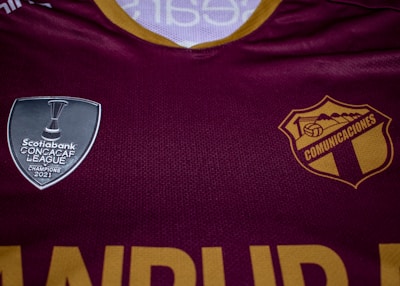 A maroon sports jersey prominently features two logos. On the left side is a silver shield with the text 'Scotiabank CONCACAF League Champions 2021'. On the right is a gold and maroon crest with 'COMUNICACIONES' printed.