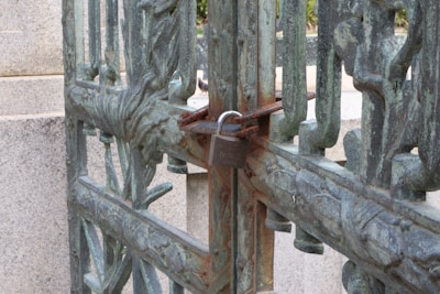 An ornate, weathered metal gate is secured with a padlock, showing intricate patterns and a rustic appearance, with stone structures in the background.