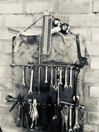 A collection of various hand tools, including wrenches, pliers, and screwdrivers, are organized neatly on a wall-mounted holder made of metal against a brick wall. The arrangement is in black and white, emphasizing the metallic texture and vintage aesthetic of the tools.