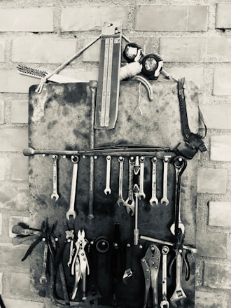 A collection of various hand tools, including wrenches, pliers, and screwdrivers, are organized neatly on a wall-mounted holder made of metal against a brick wall. The arrangement is in black and white, emphasizing the metallic texture and vintage aesthetic of the tools.