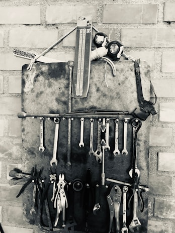 A collection of various hand tools, including wrenches, pliers, and screwdrivers, are organized neatly on a wall-mounted holder made of metal against a brick wall. The arrangement is in black and white, emphasizing the metallic texture and vintage aesthetic of the tools.