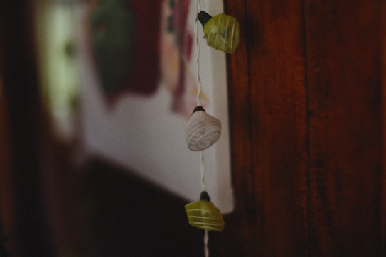 A close-up view of a string of decorative lights draped against a wooden surface. The lights are shaped like small lanterns in muted green and earthy tones. The background is blurred, suggesting an indoor setting with hints of wall art.