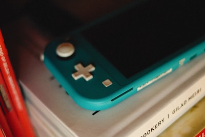 A turquoise handheld gaming console lies on top of a stack of books. The console features a white directional pad and buttons, and is partially illuminated, casting shadows across the book spines. The books are lined up vertically, with some visible text and a mix of colors including red and white.