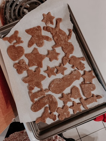 Various cutout shapes of cookie dough rest on a baking sheet lined with parchment paper, including stars, gingerbread figures, and festive shapes such as candy canes.