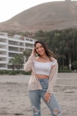 A woman stands on a sandy beach with a relaxed and confident pose. She wears a white off-the-shoulder top, light blue ripped jeans, and a beige cardigan. In the background, there are green trees and a hillside, alongside a modern white building.