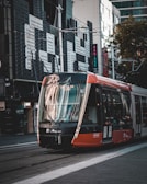 A modern tram travels on urban tracks with tall buildings and storefronts visible in the background. The tram is primarily black, red, and white and has a digital display indicating its destination as Circular Quay.