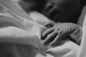 A close-up black and white photograph of a newborn baby’s hand resting on soft fabric, likely a blanket. The baby is wearing a medical wristband with partially visible numbers and text.