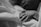 A close-up black and white photograph of a newborn baby’s hand resting on soft fabric, likely a blanket. The baby is wearing a medical wristband with partially visible numbers and text.