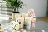 A collection of disposable paper cups is stacked on a tabletop, with some cups lying on their sides. Each cup has different designs, including logos, patterns, and text such as 'LUCKY YOU.' A potted plant is in the background, and sunlight streams through blinds, casting shadows on the table.