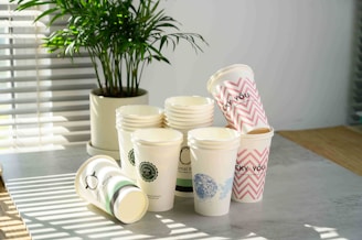 A collection of disposable paper cups is stacked on a tabletop, with some cups lying on their sides. Each cup has different designs, including logos, patterns, and text such as 'LUCKY YOU.' A potted plant is in the background, and sunlight streams through blinds, casting shadows on the table.