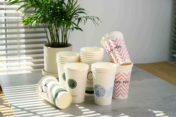 A collection of disposable paper cups is stacked on a tabletop, with some cups lying on their sides. Each cup has different designs, including logos, patterns, and text such as 'LUCKY YOU.' A potted plant is in the background, and sunlight streams through blinds, casting shadows on the table.