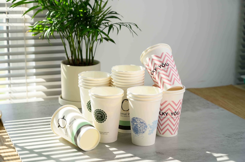 A collection of disposable paper cups is stacked on a tabletop, with some cups lying on their sides. Each cup has different designs, including logos, patterns, and text such as 'LUCKY YOU.' A potted plant is in the background, and sunlight streams through blinds, casting shadows on the table.