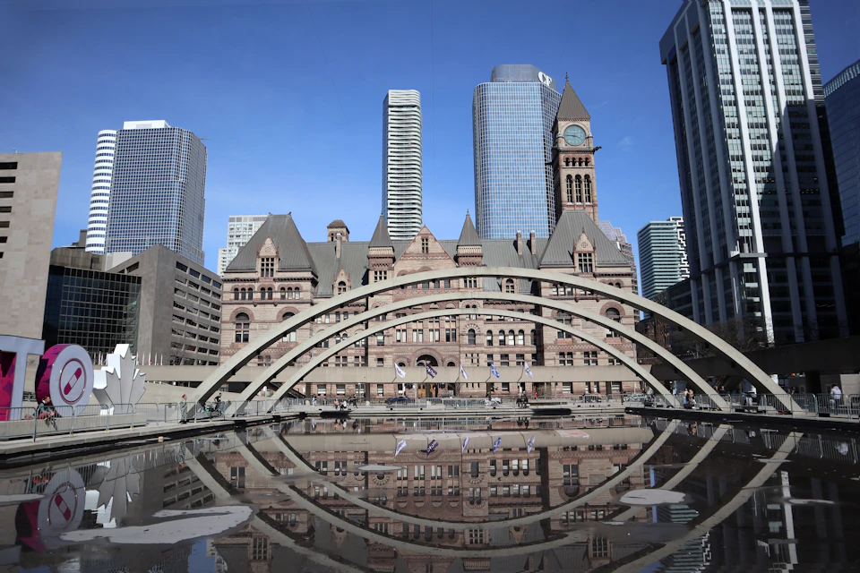 The image depicts a large, ornate building with a clock tower and multiple levels, situated on the waterfront. The building is surrounded by a large body of water that reflects its architectural details, creating a mirror-like effect. In the foreground, there are several people walking around, adding life to the scene. The sky above is clear blue, providing a beautiful backdrop for the cityscape.