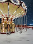 A vintage swing carousel is covered in snow at an amusement park. The ride, adorned with decorative lights and ornate details, stands still with empty seats as snowflakes fall around it. The scene is serene and captures the stillness of a winter night.
