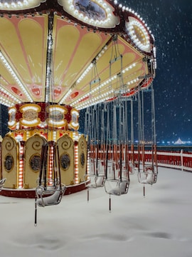 A vintage swing carousel is covered in snow at an amusement park. The ride, adorned with decorative lights and ornate details, stands still with empty seats as snowflakes fall around it. The scene is serene and captures the stillness of a winter night.