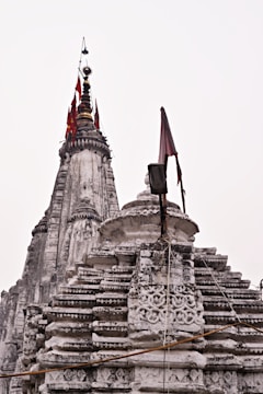 An intricately designed stone temple with ornate carvings and layered structures. The spire is topped with flags of red and gold, adding vertical contrast to the weathered stone. The architecture reflects traditional styles common in historical sites.