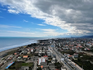 A coastal town with a dense collection of buildings spreads along the shoreline. The ocean on the left is calm, under a partly cloudy sky with patches of blue. Buildings of various sizes and colors cluster together, intersected by roads with visible traffic. Beyond the town, the landscape stretches towards distant hills.