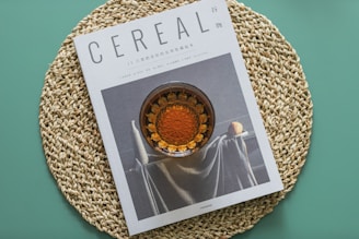 A magazine titled 'Cereal' is placed on a circular, woven placemat with a teal background. The cover of the magazine features an artistic photograph of a still life scene with a glass container filled with an amber liquid and a draped fabric beside a citrus fruit.