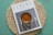 A magazine titled 'Cereal' is placed on a circular, woven placemat with a teal background. The cover of the magazine features an artistic photograph of a still life scene with a glass container filled with an amber liquid and a draped fabric beside a citrus fruit.