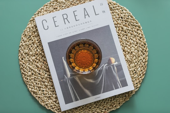 A magazine titled 'Cereal' is placed on a circular, woven placemat with a teal background. The cover of the magazine features an artistic photograph of a still life scene with a glass container filled with an amber liquid and a draped fabric beside a citrus fruit.