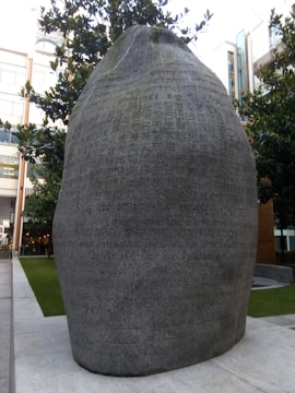 A large stone sculpture inscribed with numerous characters resembling a script. The stone is set outdoors and surrounded by trees and modern buildings, blending urban architecture with a natural element.