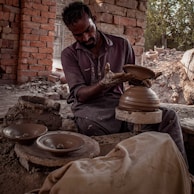 A craftsman is engaged in pottery making, shaping clay on a spinning wheel. He is surrounded by brick walls and natural lighting, with pottery pieces placed around him. His clothing appears worn with clay stains, indicating hands-on work.