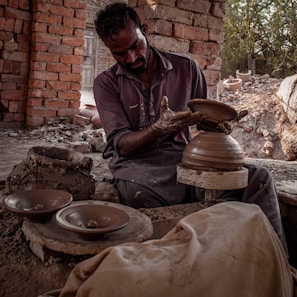 A craftsman is engaged in pottery making, shaping clay on a spinning wheel. He is surrounded by brick walls and natural lighting, with pottery pieces placed around him. His clothing appears worn with clay stains, indicating hands-on work.